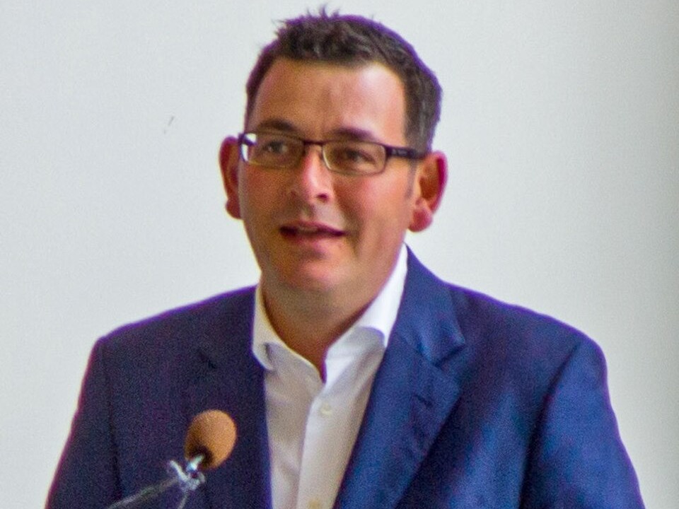 Daniel Andrews speaking at a lectern, in a blue jacket and open-necked white shirt