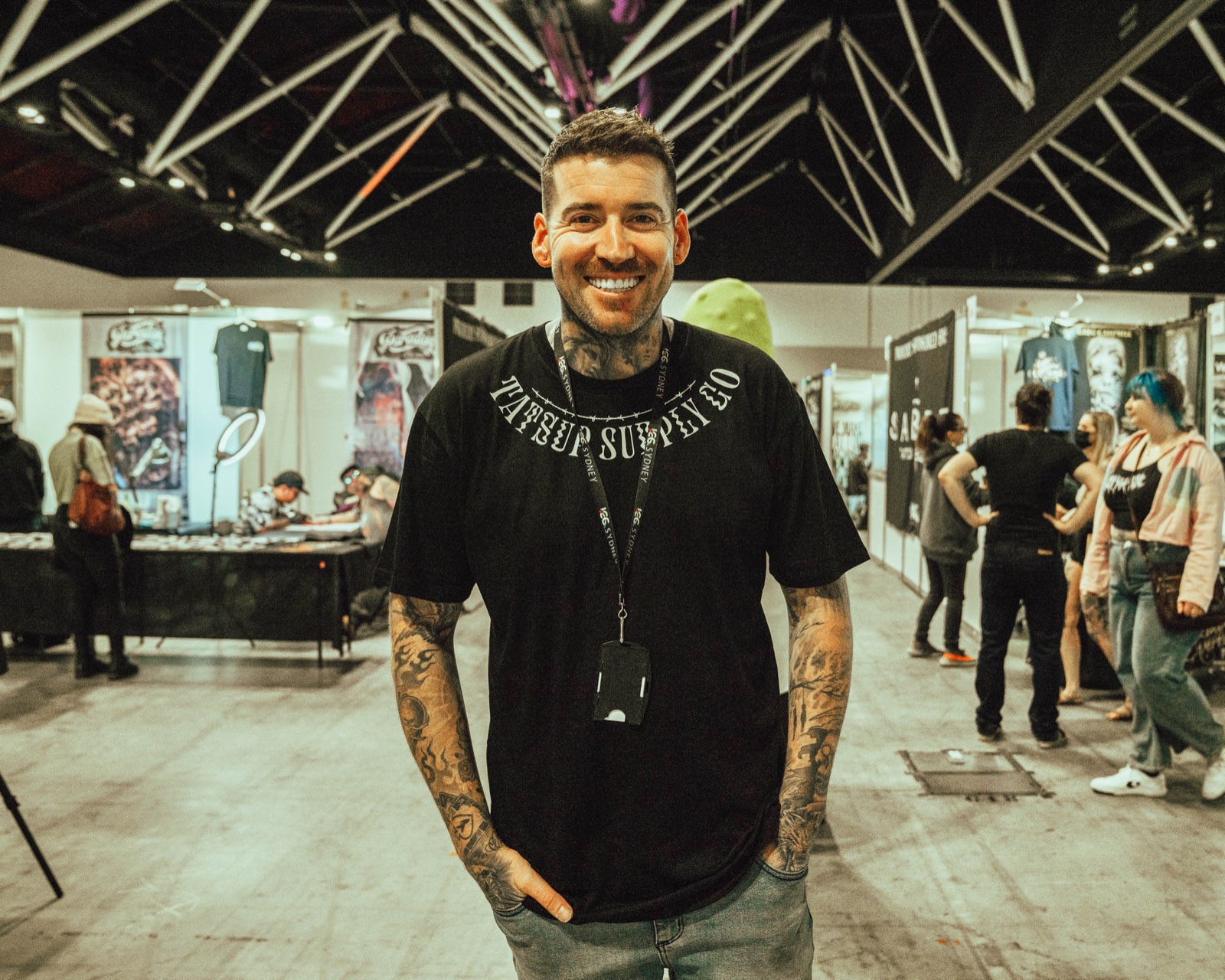 Kevin Mack standing on the floor of a tattoo expo in a Tatsup Supply Co shirt and an event lanyard, artists' booths and attendees behind him